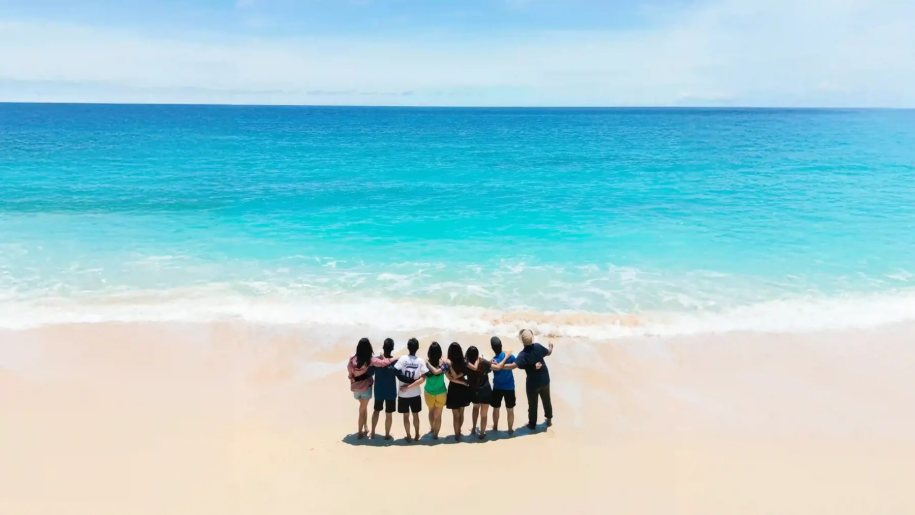 Friends arm in arm on a white sand beach facing the turquoise sea